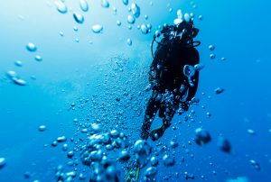 A DSLR underwater photo of a diver swimming between bubbles in the blue sea if Bora Bora, French Polynesia. The camera is aiming up at the surface and the diver is silhouetted against the bright light coming from above. The water is crispy clear blue.
